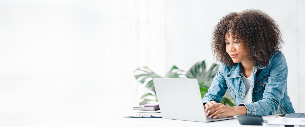 American teenage woman sitting in white office with laptop, she is a student studying online with laptop at home, university student studying online, online web education concept.