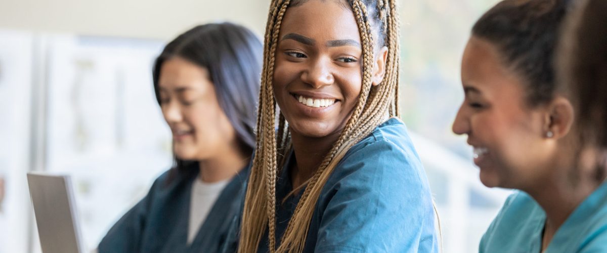 Young adult nursing or medical student wearing scrubs uses laptop computer and discusses medical training class assignment with classmate